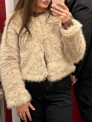 Beige fluffig pälsjacka - Säljer en supermjuk och fluffig beige pälsjacka med lång ärm och rund hals. Jackan har en kortare, boxig passform och är gjord i fuskpäls som ger en lyxig känsla. Perfekt för att hålla värmen och se stylish ut under kalla dagar.