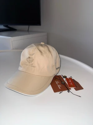 Beige keps från Loro Piana - Snygg beige keps från Loro Piana med broderad logga och emblem framtill. Klassisk böjd skärm och justerbar rem bak. Tillverkad i ett mjukt och exklusivt material som ger en clean och stilren look. Perfekt accessoar för en trendig och avslappnad stil.