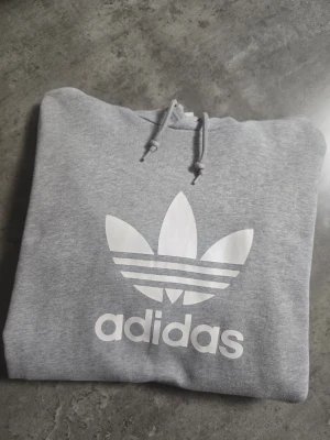 Grå Adidas hoodie med logga - Snygg grå hoodie från Adidas med stor vit Trefoil-logga och text på bröstet. Klassisk huva med dragsko och mjukt material på insidan för extra komfort. Perfekt för en avslappnad streetwear-look.