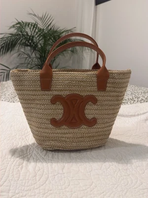Beige shoppingväska med läderdetaljer - Snygg shoppingväska i flätad beige strå med bruna läderhandtag och en stor läderdetalj framtill. Väskan har en klassisk, avslappnad form och passar perfekt för dig som gillar trendiga accessoarer med naturliga material.