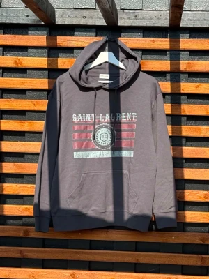 Saint Laurent hoodie  - Snygg grå hoodie med huva och dragsko. På bröstet finns ett stort Saint Laurent-tryck i vitt och rött med grafiska linjer och text. Hoodien har en klassisk känguruficka framtill och ribbade muddar vid ärmar och nederkant. Perfekt för en avslappnad streetwear-look.