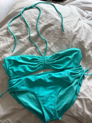 Turkos bikini med snörning, stl 36 och 38 - Snygg turkos bikini i bandeau-modell med snörning i både överdel och nederdel. Bikinin har en enkel och stilren design, perfekt för stranden eller poolen. Tillverkad i polyamid och elestan. 