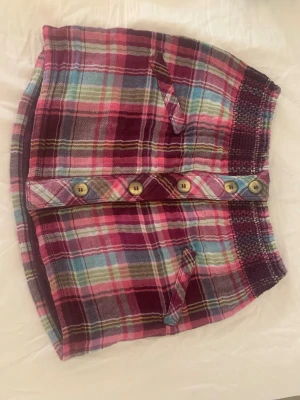 Plaid kjol - Säljer en färgglad rutig kortkjol i bomull med knappar framtill. Den har en elastisk midja och fickor som är tyvär ihop sydda