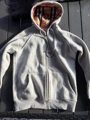 Grå zip hoodie från Burberry - Snygg grå zip hoodie från Burberry med klassiskt rutigt foder i huvan och dragsko. Hoodien har två stora fickor framtill och ribbade muddar vid ärmar och nederkant. Helt ny aldrig använd och passar bra med allt.