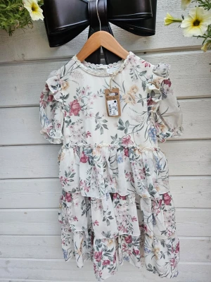 Newbie ny 128  Blommig volangklänning med puffärm - Söt klänning128  med volanger och puffärmar i ett blommigt mönster i rosa, beige och grönt på vit botten. Klänningen har rund hals med spetsdetalj och korta ärmar. Perfekt för sommarens alla tillfällen.