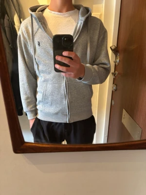 Grå zip-hoodie med broderad logga - Snygg grå hoodie med dragkedja och huva, samt dragsnören. Broderad liten logga på bröstet och två fickor framtill. Storlek m men har en lite tajtare passform. Den är nästintill oanvänd och har inga defekter