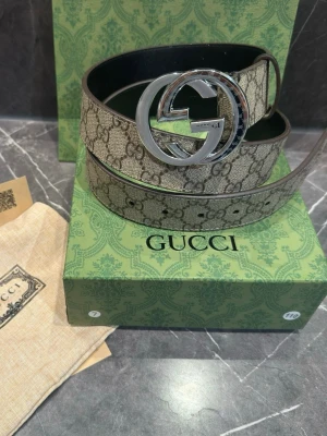 Gucci GG beige bälte med silverdetaljer - Stilrent bälte från Gucci i beige med klassiskt GG-mönster och stor silverfärgad GG-spänne. Bältet har en bred design och är tillverkat i canvas med skinninsida. Perfekt accessoar för att lyfta din outfit.