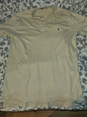 Beige pikétröja från Ralph Lauren - Klassisk beige pikétröja från Polo Ralph Lauren med blå broderad logga på bröstet. Tröjan har krage, knäppning med två knappar och är tillverkad i mjuk bomull. Perfekt för en stilren och avslappnad look.