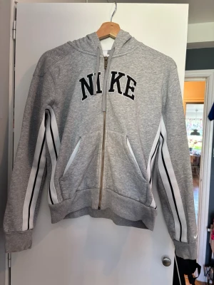 Grå zip hoodie från Nike - Snygg grå hoodie från Nike med dragkedja framtill och svartvit logga på bröstet. Har vita och svarta ränder längs ärmarna och två fickor fram. Utrustad med huva och ribbade muddar. Perfekt för en sportig och avslappnad stil.