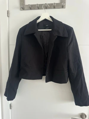 Svart kort kavaj från MNG - Stilren svart kavaj från MNG i kort modell med klassisk krage och två fickor framtill. Perfekt för att lyfta din outfit med en trendig touch. Tillverkad i ett slätt tyg med helfodrad insida för extra komfort.
