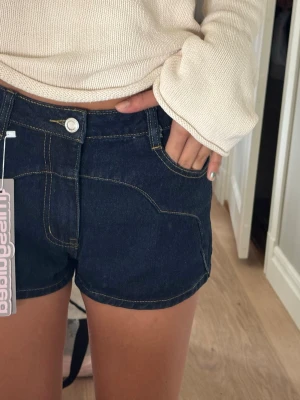 Mörkblå mini-denimshorts med kontrastsömmar - Snygga mörkblå minishorts med tydliga kontrastsömmar och klassisk femficksdesign. Shortsen har midja med knapp och dragkedja samt bälteshällor. Helt oanvända med lapp kvar!