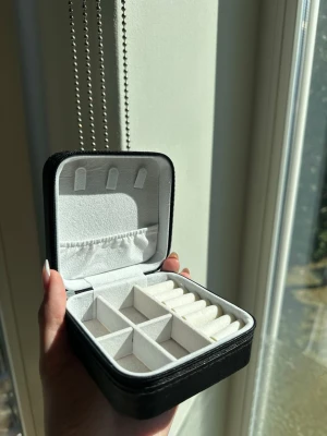 Svart smyckesbox med dragkedja - Snygg och kompakt smyckesbox i svart konstläder med dragkedja. Insidan är klädd i mjukt vitt tyg och har flera fack för ringar, örhängen och halsband. Perfekt för att organisera och ta med dina accessoarer på resan.