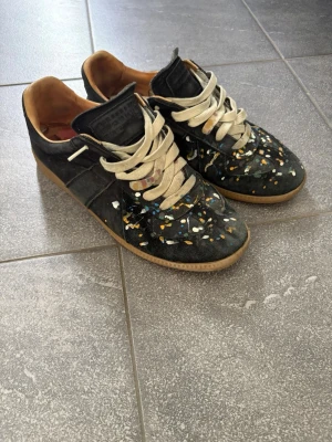 Maison margiela gats splatter  - Unika svarta sneakers med vita snören och färgstänk i blått, vitt och gult på ovandelen. Skorna har en klassisk låg profil och gummisula i beige. Insidan är ljusbrun och materialet ser ut att vara mocka eller läder.