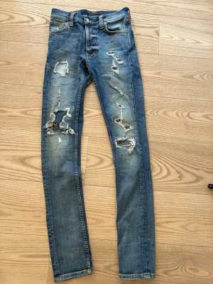 Blå slitna jeans från Nudie Jeans - Säljer ett par blå skinny jeans från Nudie Jeans med kraftiga slitningar och hål på båda benen. Jeansen har klassisk femficksmodell, orangea sömmar och är tillverkade i denim. Perfekta för en avslappnad och trendig look. De är oanvända!