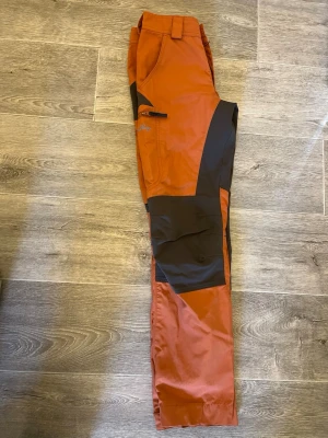 Orange och svarta friluftsbyxor från Lundhags  - 🧡 Lundhags friluftsbyxor – dam stl 34  Säljer ett par fina Lundhags friluftsbyxor i storlek 34 dam, färg orange/brun.  Byxorna är använda men är fortfarande i fint skick utan skavanker. Perfekta för vandring, friluftsliv eller andra utomhusaktiviteter. De kostar 700 kr eller bud, priset kan diskuteras🧡
