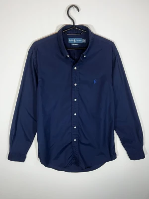 Ralph Lauren Custom Fit Skjorta Marinblå – M - Klassisk marinblå skjorta från Ralph Lauren med broderad blå Pony-logga på bröstet. Stilren och lättmatchad modell med button-down-krage.  • Storlek: M • Passform: Custom Fit • Färg: Marinblå • Broderad Polo Pony-logga • Button-down-krage • Fint begagnat skick  Passar perfekt både till vardags och mer uppklätt. 