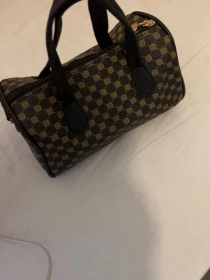 Brun mönstrad handväska med korta handtag - Snygg brun handväska med guldfärgat monogrammönster och svarta detaljer. Väskan har två korta handtag och dragkedja upptill. Klassisk boxig form och tillverkad i konstläder med textilfoder. Perfekt för dig som gillar tidlös design och vill ha plats för det viktigaste.
