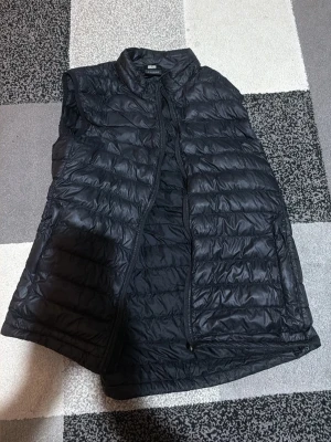 Svart dunväst från Everest - Stilren svart dunväst från Everest med quiltad design och hög krage. Västen har dragkedja framtill och är lätt vadderad för extra värme. Perfekt att slänga över en hoodie eller tröja när det är kyligt ute.