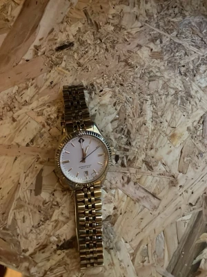 Klassisk guldfärgad armbandsklocka - Stilren armbandsklocka med guldfärgad länk och vit urtavla. Klockan har en rund boett, markerade timmarkeringar och datumvisning. Perfekt accessoar för dig som vill ha en tidlös och elegant look.