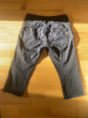 Svarta Capri jeans med knappar - Snygga svarta croppadr jeans med dekorativa knappar längs sidorna. Jeansen har två bakfickor och två framfickor, samt en knapp och dragkedja framtill. Perfekta för en avslappnad och trendig look.