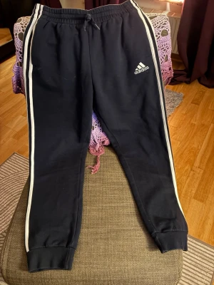 Svarta Adidas mjukisbyxor med vita ränder - Säljer ett par svarta mjukisbyxor från Adidas med klassiska vita ränder längs sidorna och Adidas-logga på låret. Byxorna har resår i midjan med dragsko och muddar vid bensluten. Perfekta för chill eller träning.