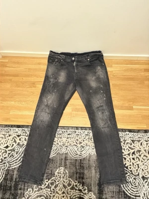 Svarta slim fit jeans med slitningar - Säljer ett par svarta slim fit jeans med coola slitningar och färgstänk på framsidan. Jeansen har fem fickor, knappgylf och en mörkröd patch bak i midjan. Perfekta för en avslappnad streetstil.