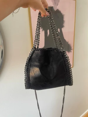 Svart handväska med kedjedetaljer - Trendig/stella McCartney liknade mörkblå handväska med grova silverfärgade kedjor som axelrem och dekor längs kanterna. Väskan har en mjuk, glansig yta och en unik form. Finns små defekter som syns på bilderna men är inget som påverkar väskans funktion 