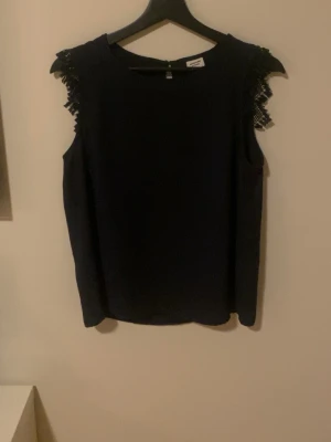 Mörkblå ärmlös topp från Jacqueline de Yong - Snygg ärmlös topp från Jacqueline de Yong med spetsdetaljer vid axlarna. Perfekt att styla med jeans eller kjol för en trendig look. Materialet känns lätt och toppen har en enkel, rak passform.