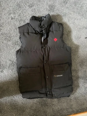 Svart dunväst från Canada Goose - Säljer en svart dunväst från Canada Goose med hög krage och två stora framfickor. Västen har en röd lönnlöv-detalj på bröstet och klassisk logotyp på insidan. Perfekt för lager-på-lager och riktigt varm under kalla dagar.