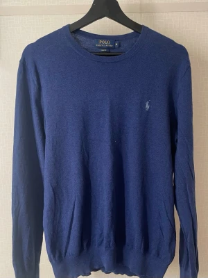Blå tröja från Polo Ralph Lauren - Klassisk blå tröja från Polo Ralph Lauren i slim fit-modell. Tröjan har rund halsringning, långa ärmar och den ikoniska broderade loggan på bröstet. Perfekt för dig som gillar stilrena och tidlösa plagg.