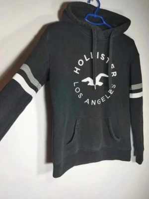 Mörkgrå Hollister hoodie med tryck - Mörkgrå hoodie från Hollister med vit logga och texten 'Los Angeles' på bröstet. Har huva med dragsko, känguruficka och två vita ränder på ärmarna. Perfekt för en avslappnad och cool stil.