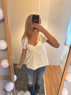 Vit blus - Söt vit blus med puffärmar och v-ringning. Toppen har en strukturerad, lite skrynklig yta och smickrande passform med rynkdetalj vid bysten. Perfekt till jeans eller kjol för en trendig look.