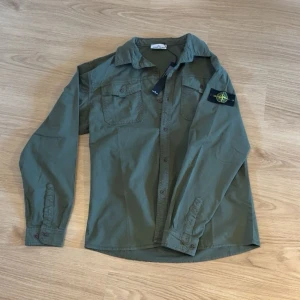 Grön OverShirt - Säljer en grön långärmad skjorta med klassisk krage och två bröstfickor. Skjortan har en ikonisk Stone Island patch på vänster ärm och stängs med knappar framtill. Perfekt för dig som gillar streetwear och vill ha en clean look.