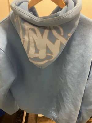 Ljusblå hoodie med vit text på huvan - Säljer en ljusblå hoodie med vita snören och stor ficka framtill. Huvan har en stor vit text i graffiti-stil på baksidan som gör hoodien unik. Perfekt för dig som vill ha en chill och snygg look.