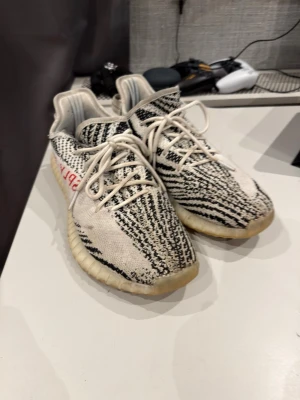 Zebra mönstrade Yeezy Boost 350 V2 - Säljer ett par Adidas Yeezy Boost 350 V2 med det ikoniska zebramönstret i svart och vitt samt texten 'SPLY-350' i rött på sidan. Skorna har en tjock, ribbad sula i beige och ovandel i stickad textil för skön passform. Perfekta för dig som gillar streetwear. Pris är diskuterbart