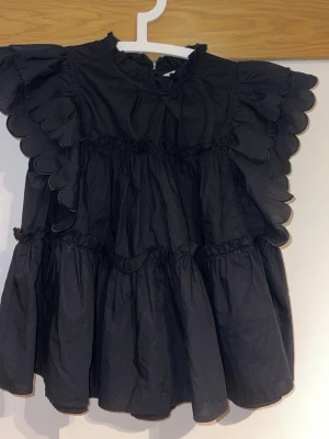 Svart volangblus med scallop-detaljer - Passar upp till 38 också. En svart blus med voluminösa volanger och scallop-kantade detaljer längs ärmarna. Blusen har hög halsringning och korta ärmar med rynkade partier.