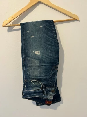 Blå slitna jeans med slimfit - Säljer ett par blå jeans med slitningar och raka ben. Jeansen har klassisk femficksdesign och detaljerade sömmar. Materialet är denim i bomull och de har en avslappnad, trendig look med distressed detaljer.