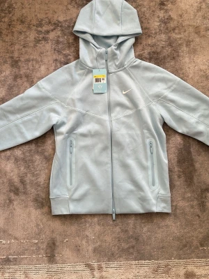 Ljusblå hoodie med dragkedja från Nike - Snygg ljusblå hoodie från Nike med hel dragkedja framtill och två fickor med dragkedja. Hoodien har en minimalistisk design med Nike-logga på bröstet och huva. Perfekt för en chill och sportig look.