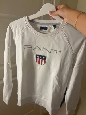 Vit sweatshirt från GANT - Vit sweatshirt från GANT med broderad logga och sköld med stjärnor och ränder på bröstet. 
