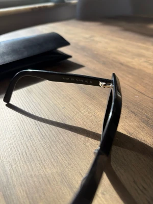 Svarta solglasögon Saint Laurent - Snygga svarta solglasögon från Saint Laurent med rektangulära bågar och mörka glas. Klassisk och stilren design som passar till det mesta. Kommer med svart fodral.