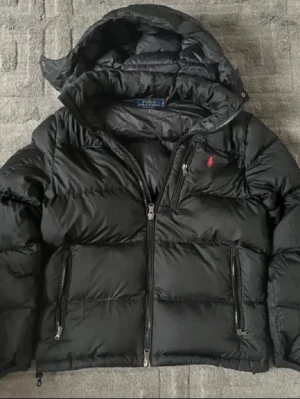 Svart Polo Ralph Lauren jacka - Säljer en svart pufferjacka från Polo Ralph Lauren med huva och två stora framfickor med dragkedja. Jackan har en röd logga broderad på bröstet och en hög krage. Perfekt för kalla vinterdagar och riktigt stilren.