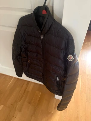 Svart dunjacka från Moncler - Snygg svart dunjacka från Moncler med quiltad design och hög krage. Jackan har dragkedja framtill, två sidofickor med dragkedja och Moncler-logga på ärmen. Perfekt för kalla vinterdagar och riktigt stilren look. Jackan är självklart äkta och är i storleken 3 som passar till S/M jackan har ett pytte litet snitt i armbågen som jag kan skicka video på privat, då det inte synd bra på bara bild 