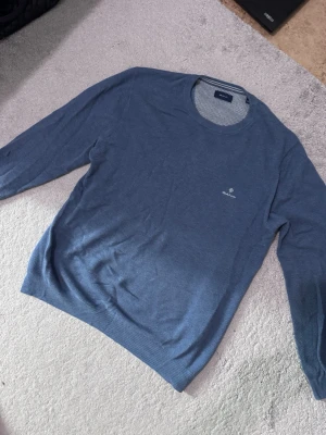 Blå stickad tröja från GANT - Snygg blå stickad tröja från GANT med rund halsringning och diskret logga broderad på bröstet. Tröjan har lång ärm och är tillverkad i ett tunt, mjukt material som passar perfekt till lager på lager. Enkel och stilren design som funkar till det mesta.