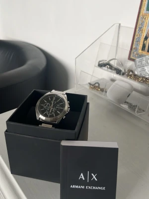 Armani Exchange silverfärgad klocka - Stilren klocka från Armani Exchange med svart urtavla och silverfärgad länk i metall. Klockan har kronograf-funktion och tydliga indexmarkeringar. Perfekt accessoar för dig som gillar en klassisk men modern look.