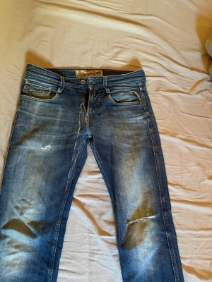 Blå slitna jeans från Replay - Säljer ett par blå jeans från Replay med slitningar och detaljerade tvättade partier. Klassisk femficksmodell med dragkedja och knapp. Jeansen har en avslappnad passform och coola slitningar på benen för en trendig look.