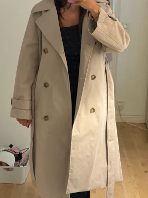 Beige trenchcoat med bälte - Stilren beige trenchcoat med dubbelknäppning och klassisk krage. Knytbälte i midjan och dekorativa knappar vid ärmsluten. Lång modell som ger en tidlös look, perfekt för lager-på-lager. Tillverkad i ett slätt tyg som känns lätt och följsamt.