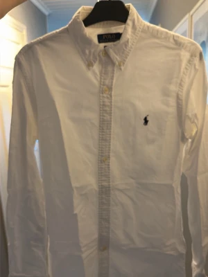 Vit skjorta från Polo Ralph Lauren - Klassisk vit skjorta från Polo Ralph Lauren i slim fit-modell. Skjortan har knappar framtill, button down-krage och den ikoniska broderade loggan på bröstet. Tillverkad i bomull och har långa ärmar. Perfekt för en clean och stilren look.