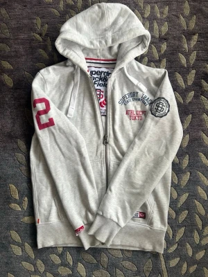 Ljusgrå Superdry hoodie med dragkedja - Ljusgrå hoodie från Superdry med ett väldigt sällsynt tryck som är väldigt svårt att får tag på. Denna zipen är prefekt nu till hösten och vintern för dig som vill hålla dig varm. Hör gärna av er om ni undrar något!!!!