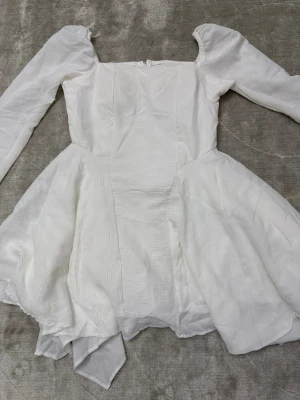 Vit långärmad kortklänning - Superfin vit kortklänning med långa ärmar och asymmetrisk volangkjol. Klänningen har en fyrkantig urringning och är gjord i ett lätt, strukturerat tyg som ger en elegant känsla. Perfekt för dig som vill sticka ut med en unik och stilren look.