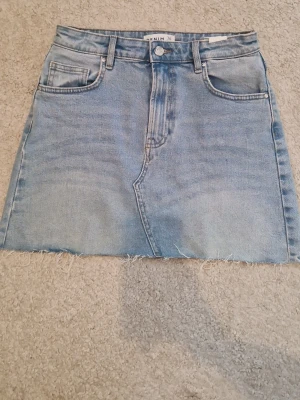 Ljusblå jeanskjol med rå kant - Säljer en ljusblå jeanskjol i klassisk denim med fem fickor och rå, fransig kant nedtill. Kjolen har normal passform och bälteshällor i midjan. Perfekt för dig som gillar en avslappnad och trendig stil.